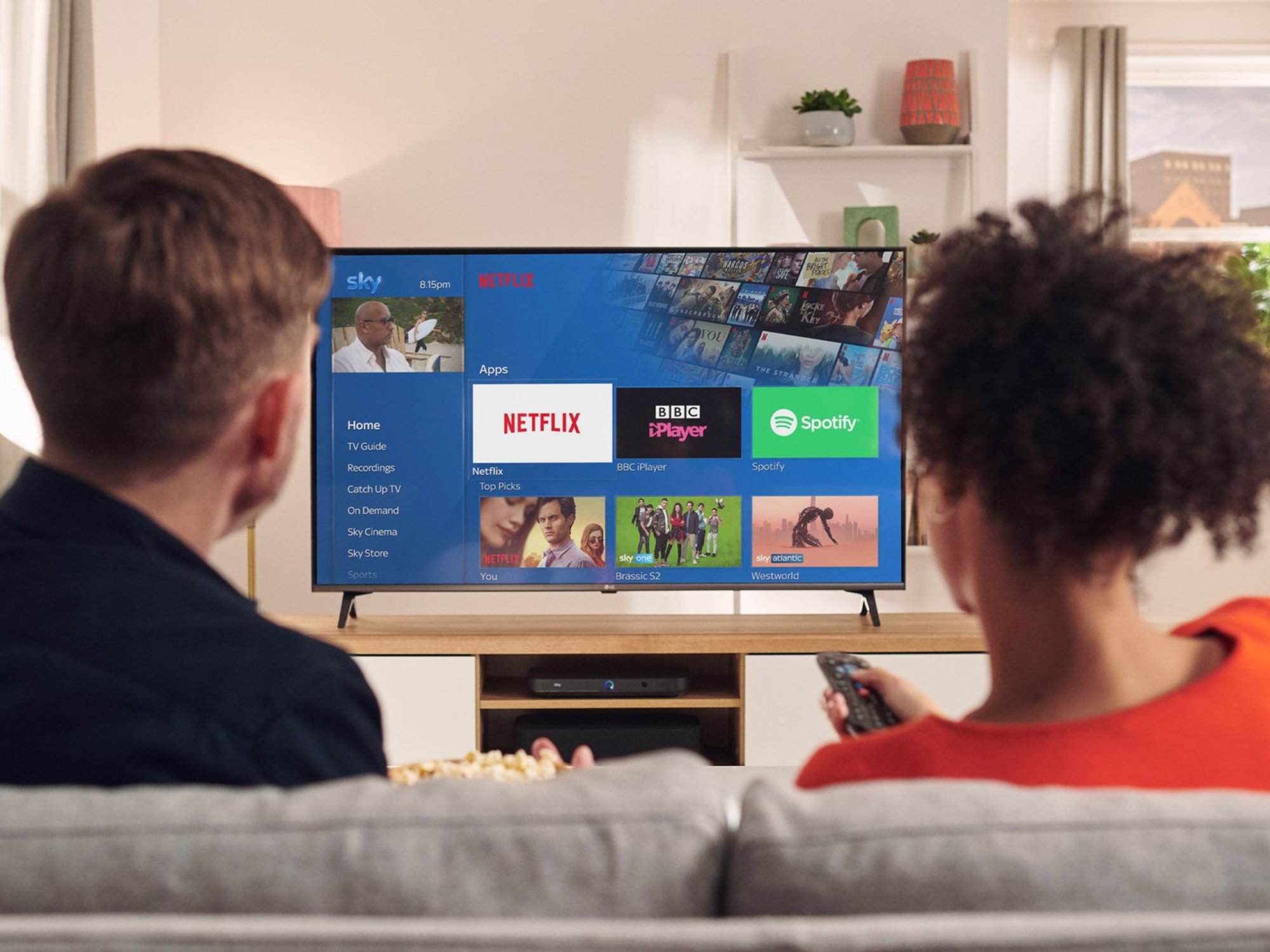 couple sits on sofa next to one another with remote in hand watching sky q box